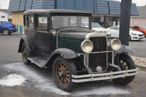 En mörk vintagebil parkerad på en asfalterad yta under lätt snöfall.